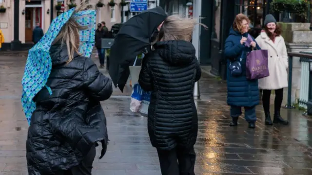 Members of the public use umbrellas to shield themselves from the wind and rain. One woman to the left of the frame is wearing a black puffer jacket and holding a patterned blue umbrella. A woman to her right is in a black jacket with a black umbrella