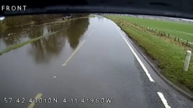 CCTV screenshot of A9 at Tomich with flood water covering most of the road. A large field of green grass is visible to the right of the frame