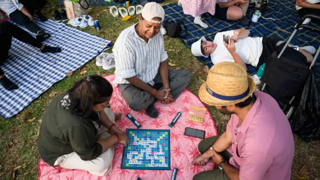 Three people sit on a pink mat surrounding a Scrabble boardgame. Two men are wearing hats and shirts while the woman is wearing a green top and beige trousers.