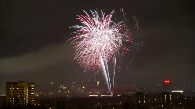 Wide shot of red, pink and green fireworks going off over Newcastle. Greyish smoke clouds to the left of the frame, buildings with turned-on lights visible from the windows at the bottom of the photo