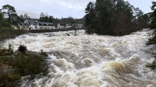 A river surging, white rapids and brown water, as a small village is behind