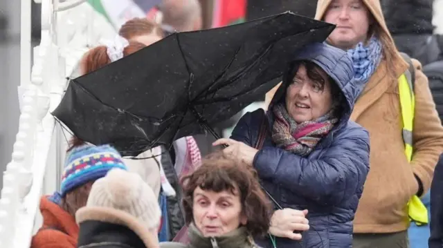 A woman in a crowd in a jacket with her hood up as it rains, battling with an inside out umbrella