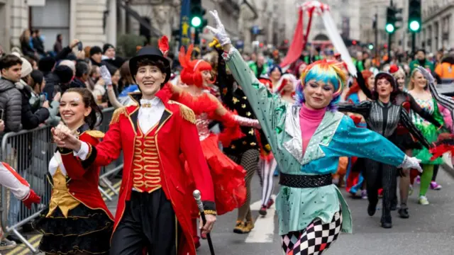 Female performers in costumes walk down Whitehall during New Year's Day Parade. A woman with short, curly brown hair dressed in a red circus master jacket with gold details and black trousers is at the centre. To her left is a woman in a multi-colours wig, hot pink turtleneck shirt, blue/silver and turquoise jacket with a glittery black belt and chequered leggins. Behind them a crowd of bystanders and other performers