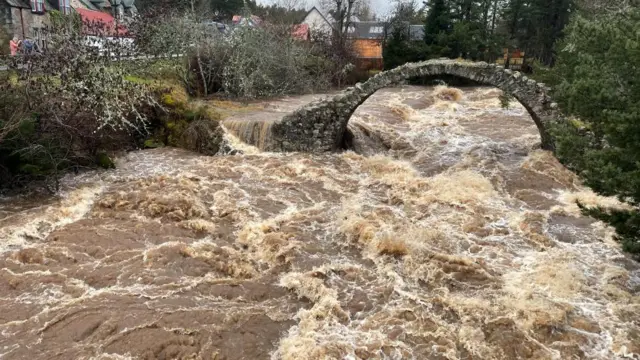 A large river is seen to be nearly overflowing, with white caps and brown mud kicked up from the bottom of the river. It's nearly reaching the bridge that crosses the river.