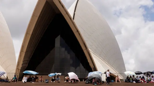 People sit in front of Sydney Opera House with towels covering their heads