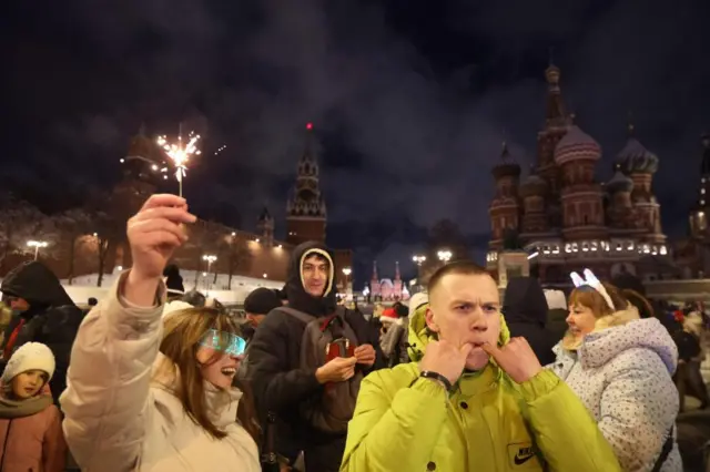 A crowd of people in front of the Kremlin buildings in the background. A man in the foreground has two fingers in his mouth, whistling. A woman next to him has a sparkler