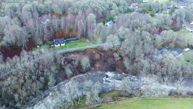 A drone shot looking down at a raging river, a huge landslide of brown mud, and houses dotted about next to a road, near the landslip