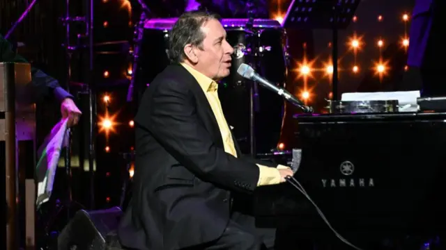 Jools Holland wearing a suit and yellow shirt playing piano and singing in front of twinkly stage lights