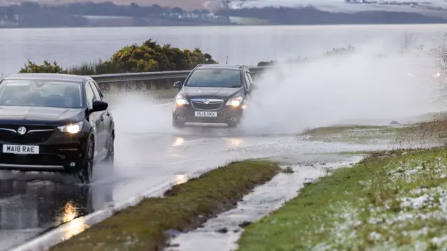 Landscape image of two large four-wheel-drive style cars driving down a road with deep puddles with water spraying up the side of the cars - a body of water is on the left and a boggy grass covered hill to the right - taken on the A9 in Inverness on 31 December