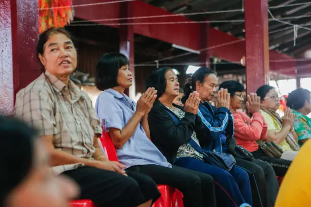 Seven women are seated on red chairs with five holding their hands in prayer looking ahead.