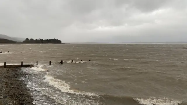 Cloudy sky over the seaside in Llanfairfechan, Conwy. Small brownish waves can be seen crashing into the pebbled beach, a medium group of trees near the bottom of a foggy hill is visible near the mid-left of the frame