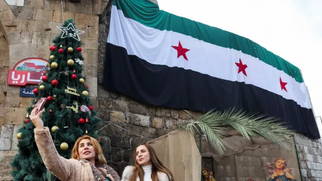 Two women pose for a selfie in front of a Christmas tree and Syrian flag