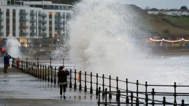 A person walking in front of a huge wave crashing in front of them as they take a picture. Another person further away does the same