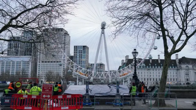 A picture of London's Eye is seen on the river with people in yellow vests working to prepare for the New Year's Eve fireworks. A sign indicates that no crossing is to take place in front of the worksite.