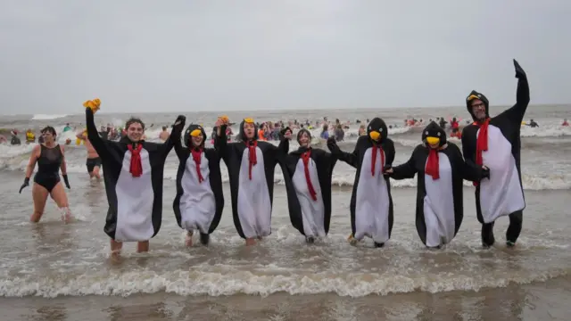 A group of seven young people dressed in penguin costumes hold hands as they celebrate taking a dip into cold water in Lyme Regis. Two men at each end of the line the group forms is holding an arm up. Beachgoers taking a dip into the greyish water