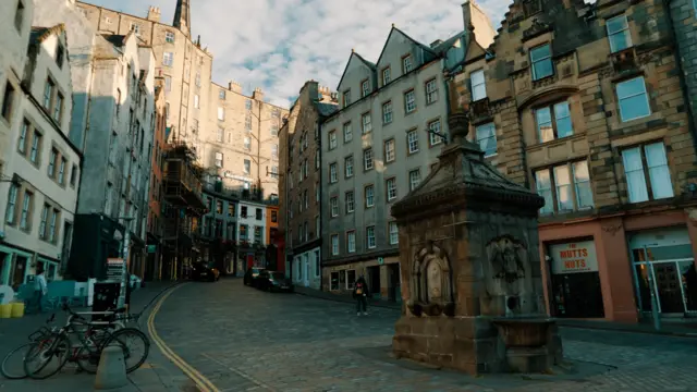 A street on a hill in Edinburgh with a big fountain in the foreground. There are no people around and the buildings are old and pretty, with sunlight on the tops of some