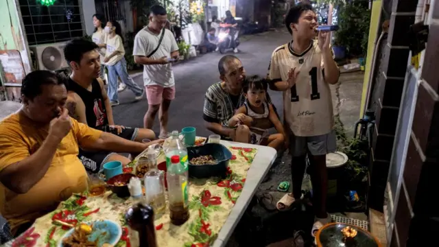 A group of men and boys gather around a table outside on a street in the Philippines. The table has food, drinks and sauces and one boy is singing karaoke.