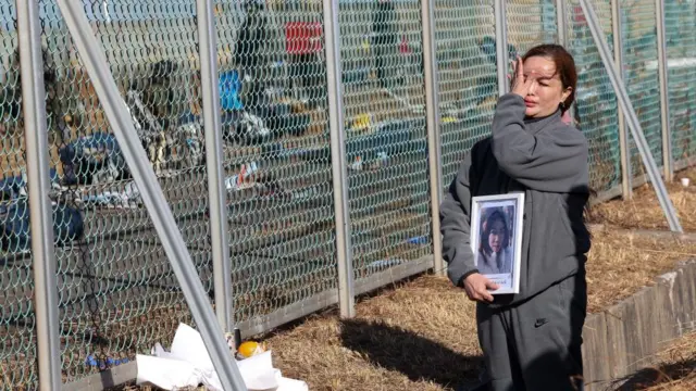 A woman holds a portrait of a person who was killed during the Jeju Air crash on Sunday. She wipes a tear away from her face as she stands in front of a fence where you can see detritus from the crash