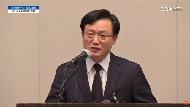 Jeju Air CEO Kim Yi-bae wearing a black suit and tie speaking into a microphone at a news conference