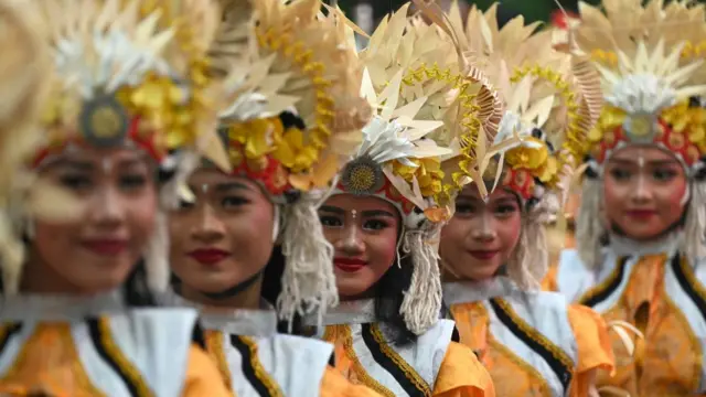 A line of traditional female dancers wearing dark eyeliner, red lipstick, and a yellow outfit with headpiece.