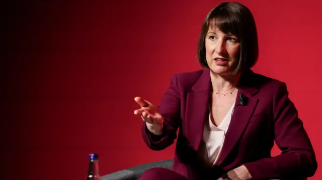 Rachel Reeves gestures as she speaks. She wears a burgundy suit and sits against a red background