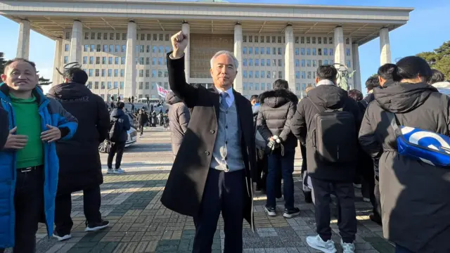 A man in a black coat holds his fist in the air outside Seoul's National Assembly building