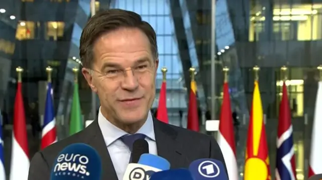 Headshot of Mark Rutte speaking into four different news microphones being held in front of him