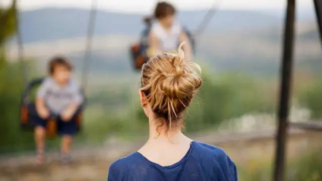 A woman stands in the foreground watching two young children playing on swings, with trees and hills in the background. The woman has her hair up and is wearing a blue top.