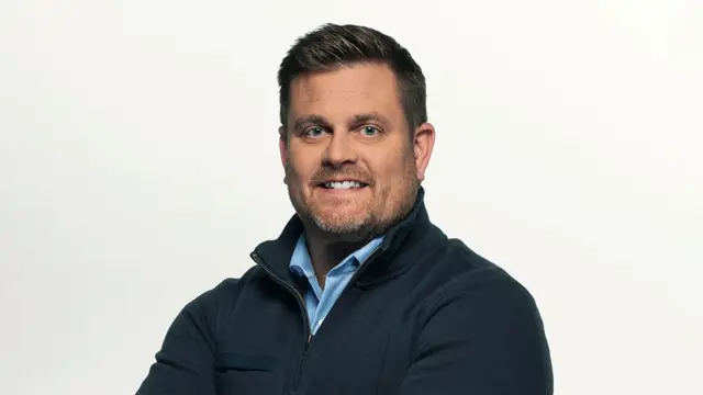 Brian Thompson looks into the camera, smiling and wearing a blue collared shirt and sweater, against a white backdrop in a headshot