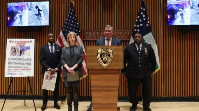 Four people stand in a line, between two flags and two television screens showing CCTV footage. One man, the third along in the line, is wearing a navy suit and is behind a podium, speaking into a microphone. The rest are all wearing dark formal clothing