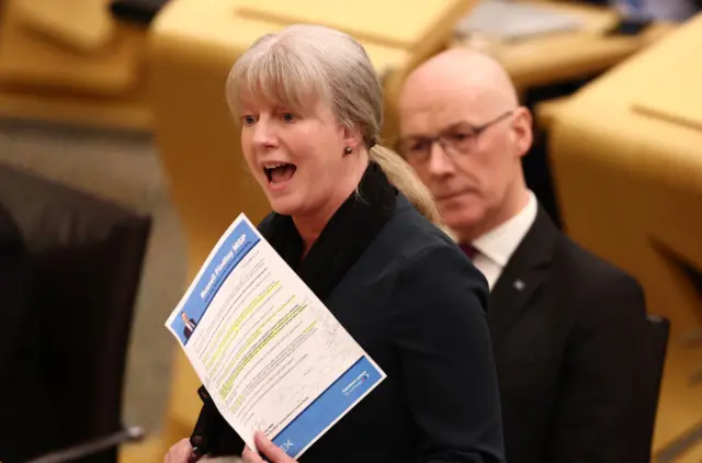 Shona Robison speaking in the parliament while waving a piece of paper. John Swinney is sitting behind her.