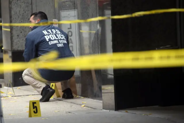 A NYPD officer crouches outside the Hilton Hotel where a shooting took place. There is yellow police tape in front of him