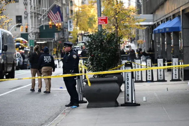 Wide shot showing outside of Hilton hotel with yellow tape marking crime scene. Several officers at the scene. American flags on a building opposite the road.