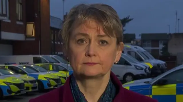 Headshot of Yvette Cooper. There's a row of police cars and vans behind her