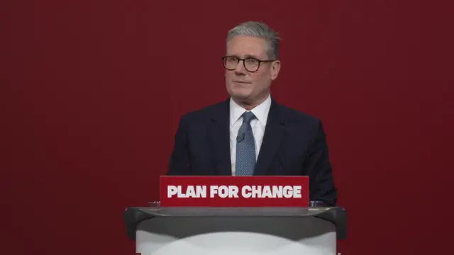 Keir Starmer, wearing a suit, answers questions from journalists behind a podium with a sign reading Plan for Change