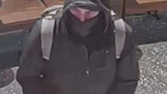 A still from security footage shows a  person is wearing a black jacket, a black balaclava covering the lower half of their face, and a grey backpack