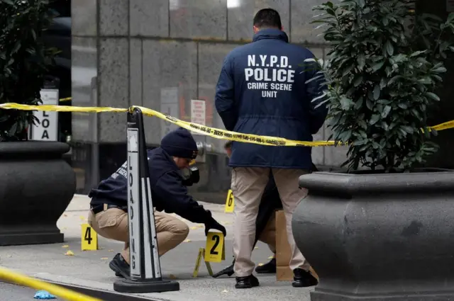 Crime scene outside Manhattan's Hilton Hotel with yellow tape around the scene. Two officers with jackets labelled NYPD police Crime Scene Unit, one is standing, the other is placing a numbered yellow card on the ground