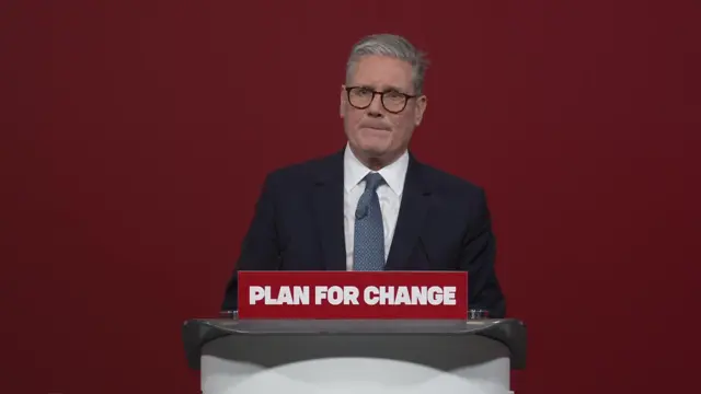 Keir Starmer answers questions from journalists in a suit standing behind a podium with a sign reading 'Plan for Change'