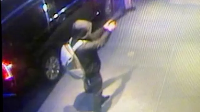 Blurry CCTV image of the suspected gunman, shot from the back with a side profile, standing on a path, with arms holding a gun and pointing to the right with a parked car to his left. He's wearing a dark hoodie, with a grey backback.