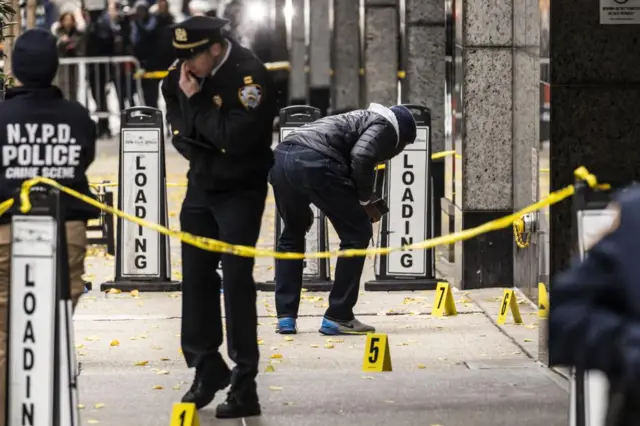 Crime scene outside Manhattan's Hilton Hotel with yellow tape around the scene. Three people within the cordon. One wearing a NYPD Police Crime Scene unit jacket, another in uniform and another in casual clothes bending down to inspect evidence on the ground. Several numbered yellow cards are on the ground to mark evidence