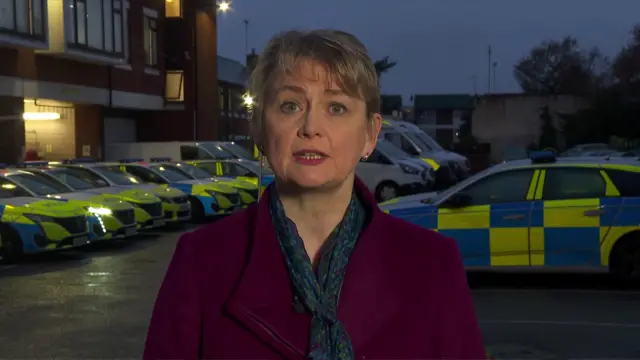 Yvette Cooper wearing a maroon coat and standing in a car park, with police cars in the background