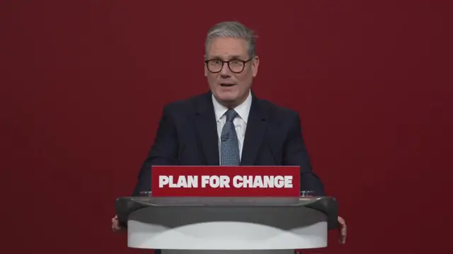 Keir Starmer answers questions from journalists standing behind a podium with a sign reading Plan for Change