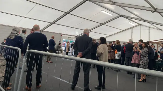 A photo taken from behind a fence of people standing about inside a white tent