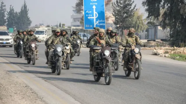 Several men riding motorbikes in camouflage army fatigues. A blue road sign with directions to Hama and Damascus - signposted in both English and Arabic - is behind them.