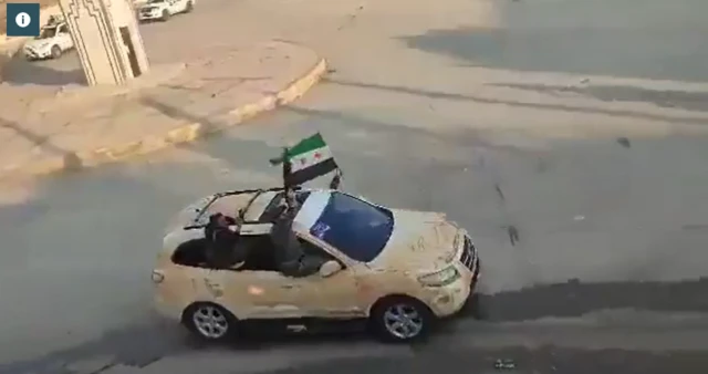 Ariel image of a light brown car with two men hanging out of the near side. A black, green and white flag representing the Syrian opposition is flown out of a far side window