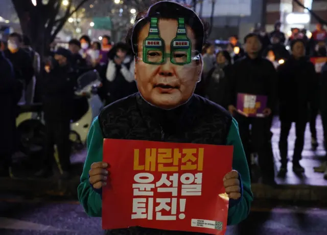 A protester wearing a mask showing the face of President Yoon Suk Yeol holds a placard reading, "Yoon Suk Yeol, step down for treason!"