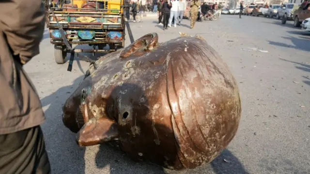 A large statue head badly damaged with what appear to be bullet holes is dragged along a street behind a vehicle