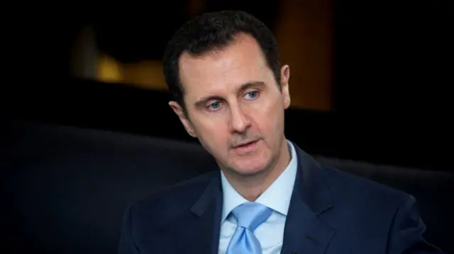 Bashar al-Assad mid shot cutting off below shoulders. He's sitting down in dark blue suit, light blue shirt and tie. Background is dark and blurred