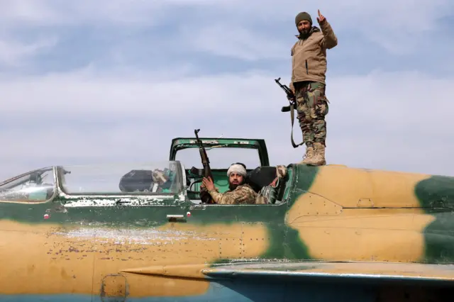 Two Syrian rebel forces onboard a Syrian army jet, one inside the cockpit, holding rifles.