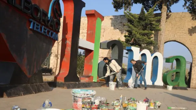 Two men paint over the letters of a large "Hama" sign